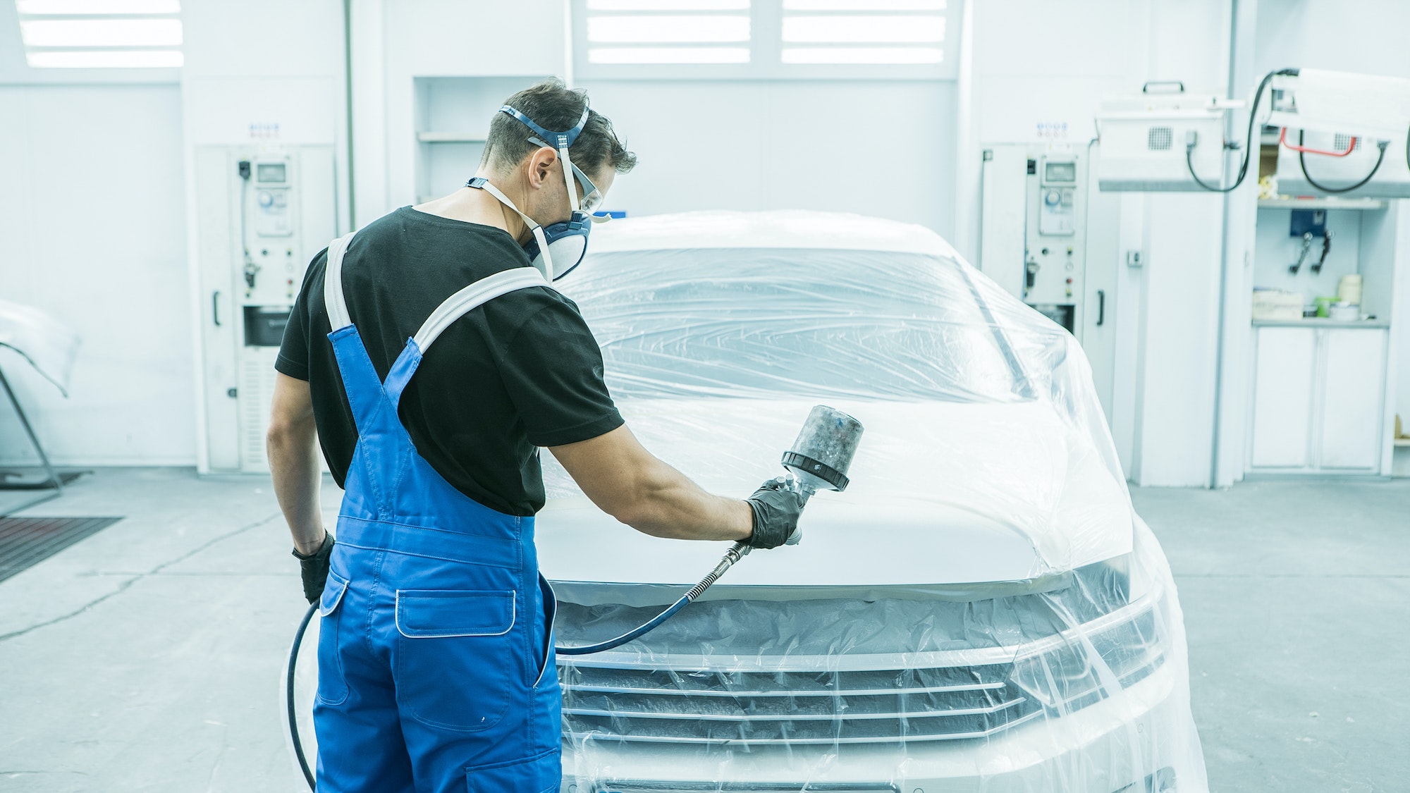 A car painter wearing protective clothing paints a car with an airbrush in an auto repair shop.